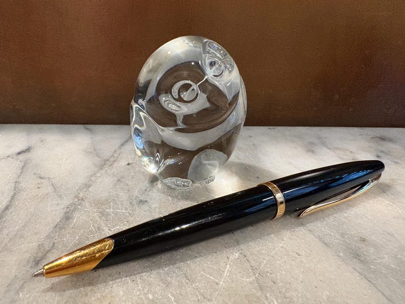 Puede incluir: Un bol&iacute;grafo negro con detalles en color dorado y una punta dorada, colocado frente a una figura de b&uacute;ho de cristal macizo transparente. El bol&iacute;grafo presenta una banda dorada alrededor del centro y un clip dorado en la tapa.