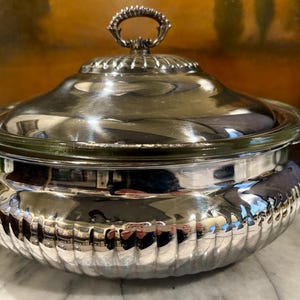May include: A silver-plated serving dish with a lid and a handle. The dish has a ribbed design around the base and a reflective surface. The lid has a decorative handle. The dish is likely used for serving food.