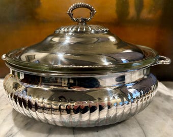 May include: A silver-plated serving dish with a lid and a handle. The dish has a ribbed design around the base and a reflective surface. The lid has a decorative handle. The dish is likely used for serving food.