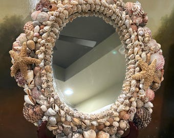May include: Oval mirror with a frame entirely covered in various seashells and starfish. The seashells are in shades of beige, brown, and white. The mirror is supported by a dark brown wooden stand, creating a coastal or nautical theme.