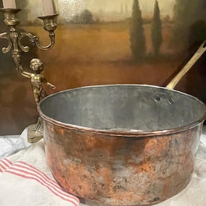 May include: A large, antique copper pot with a brass handle. The pot has a worn, aged patina and is sitting on a white surface.