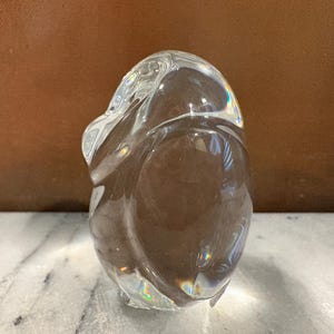 Puede incluir: Una escultura de vidrio s&oacute;lido transparente con forma de ping&uuml;ino estilizado. El objeto presenta contornos suaves y redondeados, con una superficie pulida y reflectante que capta la luz, creando sutiles refracciones prism&aacute;ticas en el material.