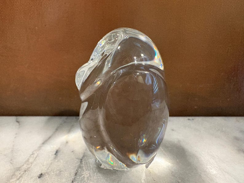 Puede incluir: Una escultura de vidrio s&oacute;lido transparente con forma de ping&uuml;ino estilizado. El objeto presenta contornos suaves y redondeados, con una superficie pulida y reflectante que capta la luz, creando sutiles refracciones prism&aacute;ticas en el material.