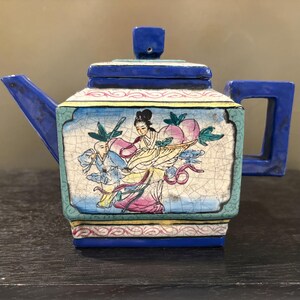 May include: A square ceramic teapot with a deep blue lid, handle, and spout. The body features a crackled cream-colored panel depicting a figure in traditional robes holding a large peach, alongside a smaller figure, framed by teal and pink borders.