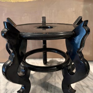 May include: A black wooden display stand featuring a circular top platform with a central hole, supported by four curved, ornate legs that extend outward. The stand has a smooth, polished finish with visible signs of wear along the edges.