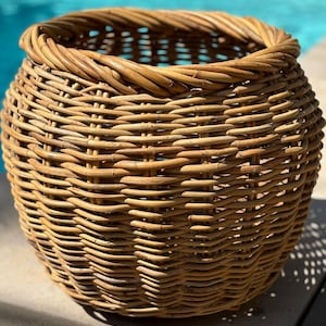 May include: A round, woven wicker basket with a natural, light brown color. The basket has a wide opening and a sturdy, rolled rim. The weave pattern is tight and consistent, creating a textured surface. The basket is sitting on a light-colored surface.