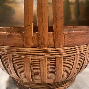 Puede incluir: Una cesta de bamb&uacute; marr&oacute;n tejida con asa. La cesta tiene una escena pintada de un paisaje con &aacute;rboles y una masa de agua.