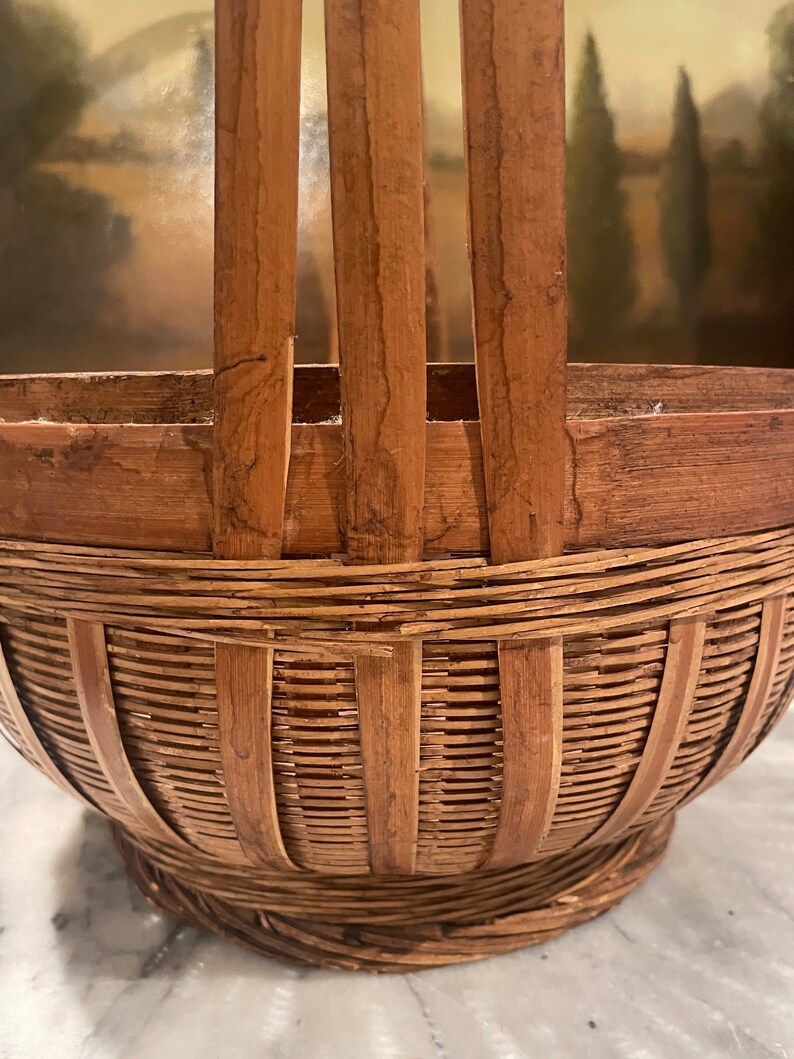 Puede incluir: Una cesta de bamb&uacute; marr&oacute;n tejida con asa. La cesta tiene una escena pintada de un paisaje con &aacute;rboles y una masa de agua.