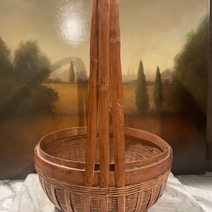 Puede incluir: Una cesta de bamb&uacute; tejida grande, de color marr&oacute;n, con un asa alta. La cesta est&aacute; hecha de materiales naturales y tiene un aspecto r&uacute;stico y vintage.