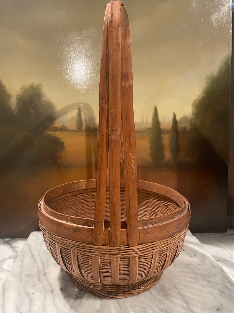 Puede incluir: Una cesta de bamb&uacute; tejida grande, de color marr&oacute;n, con un asa alta. La cesta est&aacute; hecha de materiales naturales y tiene un aspecto r&uacute;stico y vintage.