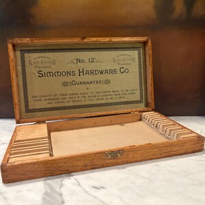 May include: A rectangular wooden storage box with a hinged lid featuring a printed label that reads Simmons Hardware Co. No. 12 Guarantee. The interior contains molded slots for organizing items, with a light-colored base and a metal latch closure.