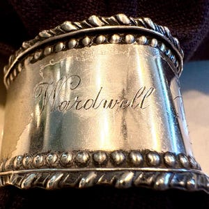 May include: A silver-toned metal napkin ring featuring a polished center band engraved with the name Wardwell in cursive script. The top and bottom edges are decorated with a beaded border and a twisted rope-like trim, encircling a dark fabric.