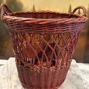 May include: A tall, reddish-brown wicker basket with two curved handles. The basket features a woven design with a lattice pattern on the sides and a solid woven base. The basket is likely used for storage or decoration.