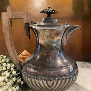 May include: A silver teapot with a wooden handle and a decorative design. The teapot has a black knob on the lid.
