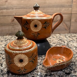 May include: A brown clay tea set featuring a teapot with a lid, a lidded sugar bowl, and a small open creamer. Each piece is decorated with hand-painted white and black floral patterns and intricate border designs on the rims and lids.
