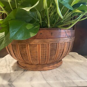 Puede incluir: Una cesta redonda de mimbre marr&oacute;n con un borde de madera, llena de una planta verde de hojas. La cesta tiene una base tejida y est&aacute; sentada sobre una superficie de m&aacute;rmol blanco.