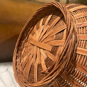 Puede incluir: Una cesta de mimbre marr&oacute;n con una parte superior redonda y abierta y un patr&oacute;n tejido. La cesta est&aacute; hecha de materiales naturales y tiene un aspecto r&uacute;stico.