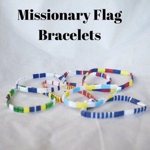 May include: A group of colorful bracelets with a white background. The bracelets are made of small, square beads in red, white, blue, yellow, and green. The bracelets are arranged in a loose circle, with some overlapping. The text "Missionary Flag Bracelets" is written in black letters at the top of the image.