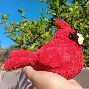 May include: A bright red crocheted cardinal bird plush toy with a soft, textured yarn finish. The bird features a distinct black patch around its small, light-colored beak and dark, round eyes, showcasing a detailed, handcrafted design.