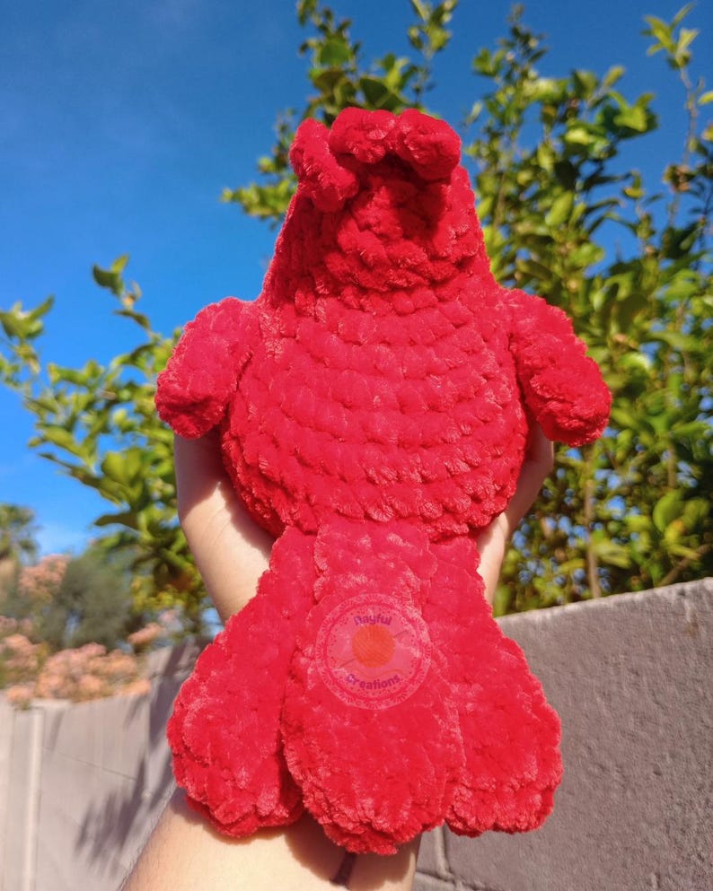 May include: A bright red plush bird toy crafted from soft, textured yarn. The bird features a rounded body, small wings, and a prominent tail. A circular logo with the text Playful Creations is visible on the lower section of the tail feathers.