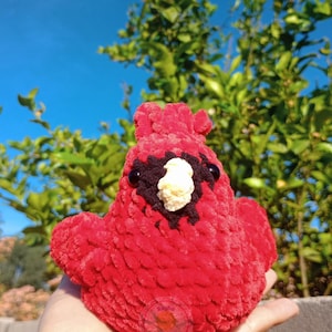 May include: A bright red crocheted cardinal bird plush toy with a soft, fuzzy texture. It features a black mask around the eyes, a small white beak, and two dark, round plastic eyes. The bird has a rounded body shape with small wings.