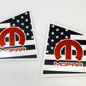 May include: Two decals with a design based on the American flag, featuring a red Mopar logo. The flag has black and white stripes and a field of white stars. The word "MOPAR" is printed in red.