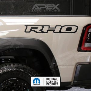 Pode incluir: Uma vista de perto de uma cama de caminhão bege com um decalque "RHO" em preto e branco. O caminhão tem um pneu preto e um pára-lama preto. O logotipo "OFFICIAL LICENSED PRODUCT" é visível.
