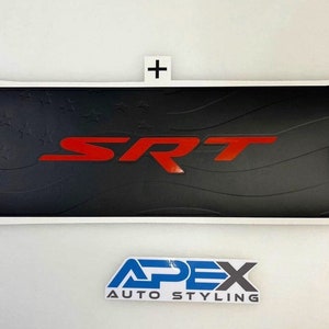 Può includere: Accessorio per auto in bianco e nero con le lettere rosse "SRT" al centro. L'accessorio presenta un disegno di stelle e strisce. Il testo "APEX AUTO STYLING" è in basso.