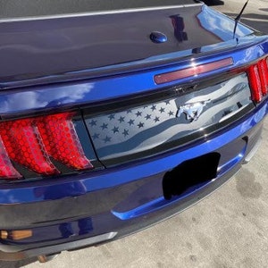 May include: A blue sports car's rear with red taillights. The car features a black panel with an American flag design and a silver Mustang emblem. The taillights have a honeycomb pattern.