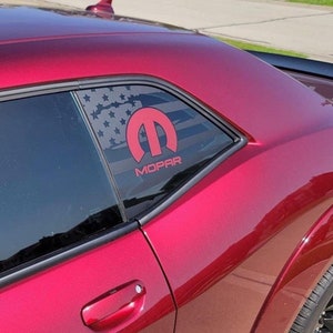 Puede incluir: Un coche rojo con una calcomanía en la ventana que muestra la bandera estadounidense y el logotipo de Mopar en rojo. La pintura del coche tiene un acabado brillante y la imagen está tomada al aire libre a la luz del día.