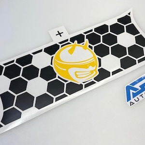 May include: A white sticker with a black and white honeycomb pattern and a yellow cartoon character. The sticker has a plus sign on a small white tab. A smaller sticker with blue text is also present.