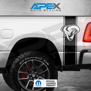 Puede incluir: Parte trasera de una camioneta blanca con franjas negras y un gráfico de una cabeza de carnero. El texto "APEX AUTO STYLING" está en la parte superior. El neumático dice "All-Terrain T/A". El logotipo del producto con licencia oficial es visible.