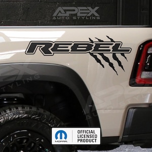 May include: A tan truck bed with a black "REBEL" decal and claw marks. The decal is outlined in white. The truck has a black tire and fender. The image also includes the "OFFICIAL LICENSED PRODUCT" logo.