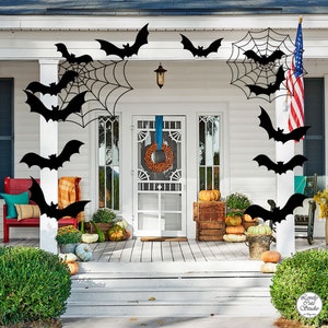 May include: A white house with a black front door and a wreath on it. There are black bats and spiderwebs hanging from the eaves. There are pumpkins and gourds on the porch.