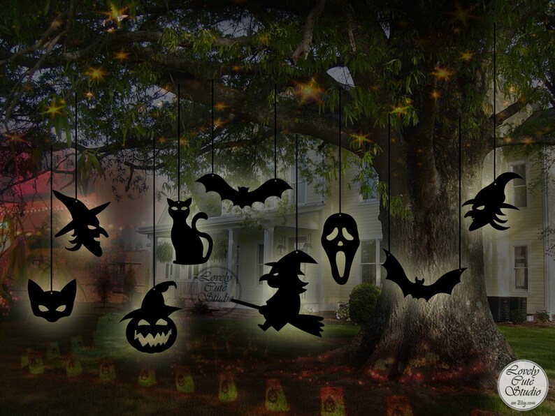 May include: Black silhouette cutouts of Halloween characters hanging from a tree branch. The characters include a witch, a cat, a bat, a ghost, a pumpkin, and a skull. The cutouts are illuminated by string lights.