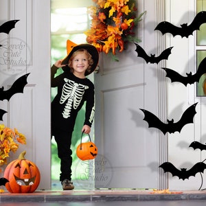 May include: A child dressed as a skeleton in a black costume and a witch hat holds a pumpkin bucket. The child is standing in a doorway decorated with black bats and a spiderweb. There are pumpkins and fall foliage on the porch.