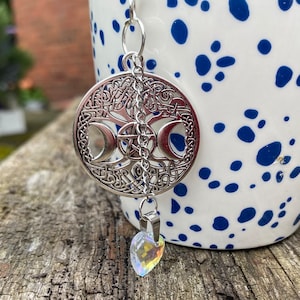 Puede incluir: Colgante plateado con diseño de triple luna, pentáculo y nudos celtas. Un pequeño corazón de cristal iridiscente cuelga debajo. El colgante está suspendido por una cadena. Fondo: taza blanca con lunares azules y superficie de madera.