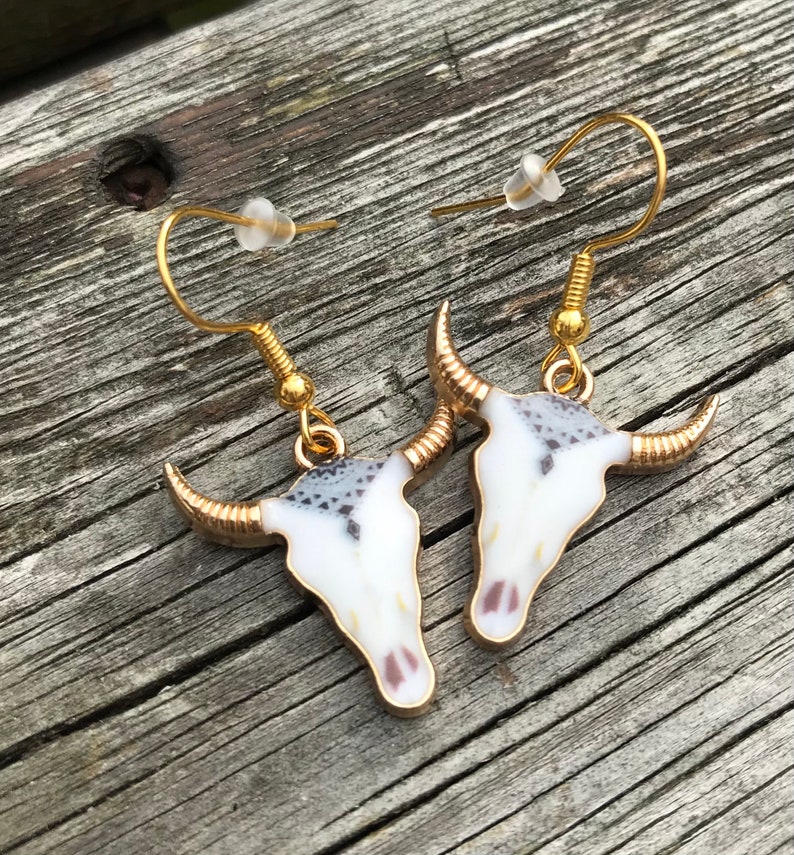 K&ouml;nnte beinhalten: Ein Paar goldfarbene Ohrringe mit einem wei&szlig;en und grauen Totenkopfdesign. Der Totenkopf hat goldene Akzente an den H&ouml;rnern und einen wei&szlig;en Hintergrund mit grauen und schwarzen Details.