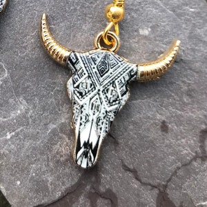 May include: A pair of gold and black bull skull earrings with a geometric pattern. The earrings are made of metal and have a hook closure.