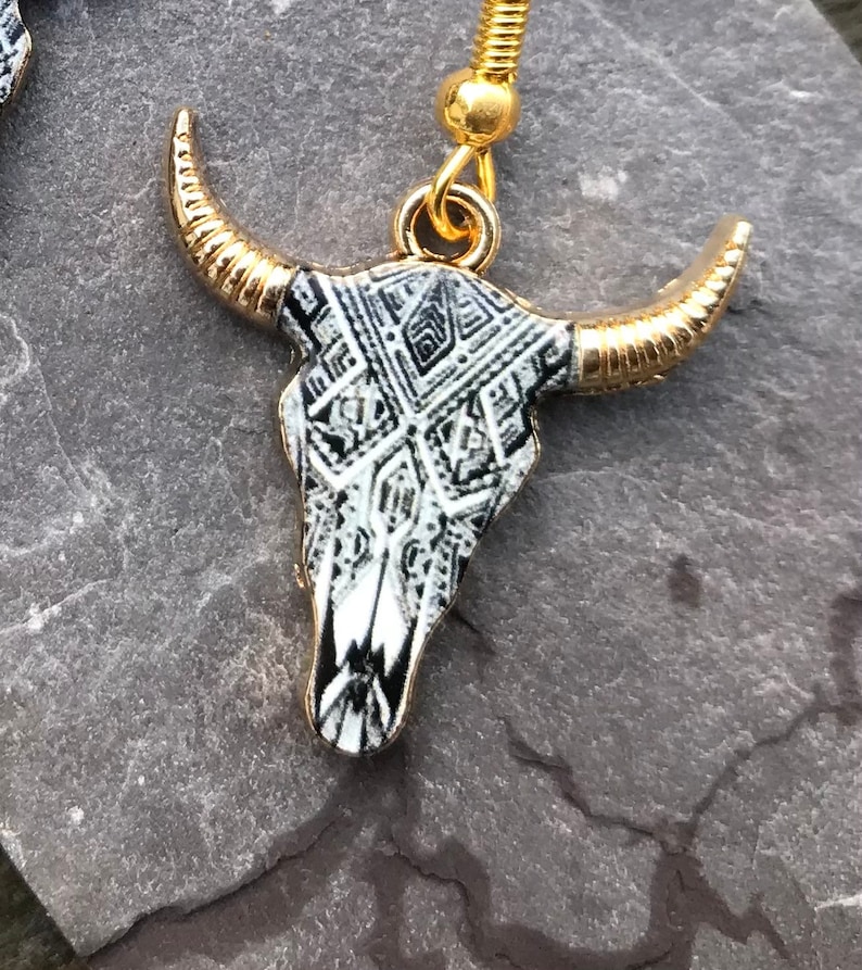 May include: A pair of gold and black bull skull earrings with a geometric pattern. The earrings are made of metal and have a hook closure.