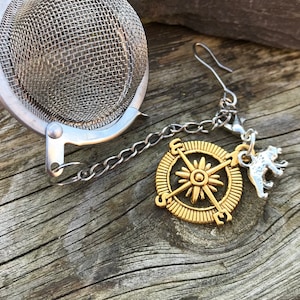 Puede incluir: Un infusor de té de plata con una cadena unida a un dije de brújula de oro y un dije de oso de plata.