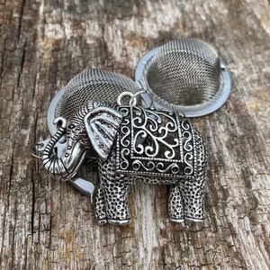 Op de afbeelding: Zilveren thee-ei in de vorm van een olifant met ingewikkelde details. De thee-ei is open aan de boven- en onderkant, waardoor theebladeren erin kunnen worden geplaatst. De thee-ei ligt op een houten oppervlak.