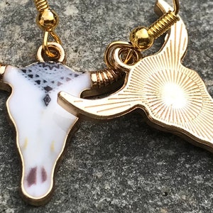 K&ouml;nnte beinhalten: Ein Paar goldfarbene Ohrringe mit einem Stierkopfdesign. Ein Ohrring hat einen wei&szlig;en Emaille-Sch&auml;del mit schwarzen und grauen Details, w&auml;hrend der andere Ohrring ein massiver goldfarbener Sch&auml;del mit einem Sonnenstrahlendesign ist.