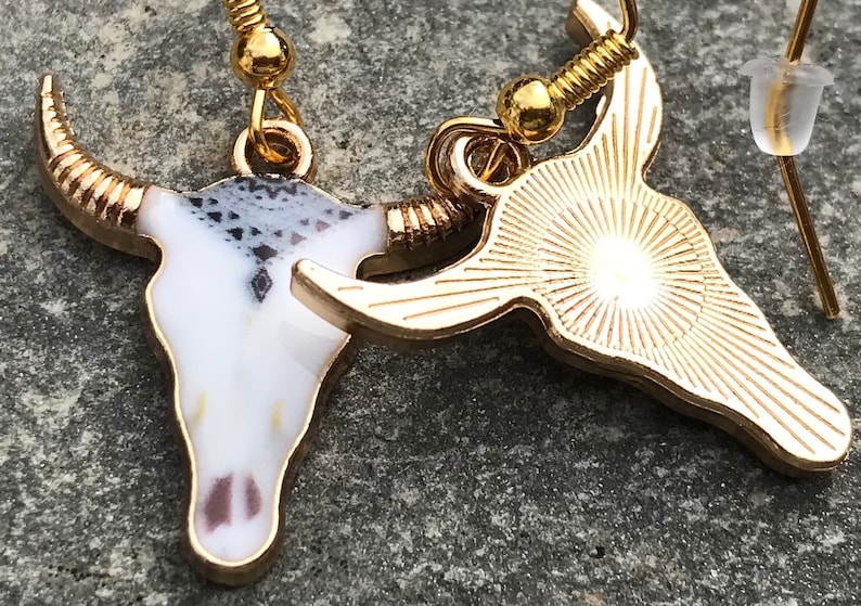 K&ouml;nnte beinhalten: Ein Paar goldfarbene Ohrringe mit einem Stierkopfdesign. Ein Ohrring hat einen wei&szlig;en Emaille-Sch&auml;del mit schwarzen und grauen Details, w&auml;hrend der andere Ohrring ein massiver goldfarbener Sch&auml;del mit einem Sonnenstrahlendesign ist.
