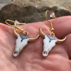 K&ouml;nnte beinhalten: Ein Paar goldfarbene Ohrringe mit einem wei&szlig;en und schwarzen Stierkopfdesign.