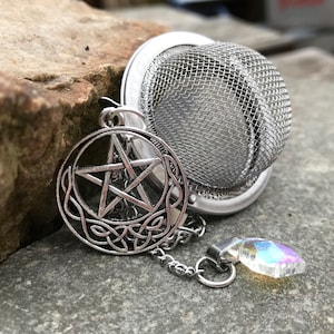 Könnte beinhalten: Silberner Tee-Ei mit einem Pentagramm-Anhänger und einem Kristall-Anhänger. Der Tee-Ei ist kugelförmig mit einem Netzdeckel. Der Pentagramm-Anhänger ist an einer Kette befestigt.