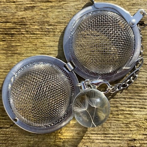 May include: A silver tea infuser with a chain and a clear glass ball charm containing a dandelion. The infuser has a mesh design and is open to show the inside.