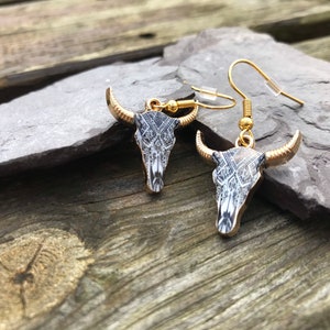 May include: A pair of gold tone earrings with a black and white patterned bull skull design.