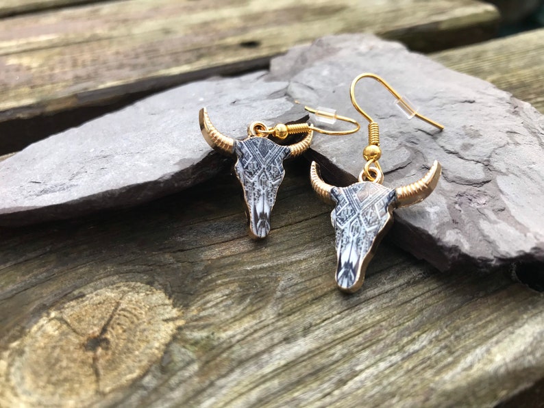 May include: A pair of gold tone earrings with a black and white patterned bull skull design.