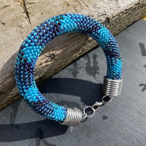 Pode incluir: Uma pulseira artesanal com um design tecido em tons de azul e preto. A pulseira apresenta extremidades de metal prateado e um fecho. O diâmetro é de aproximadamente 7 cm.